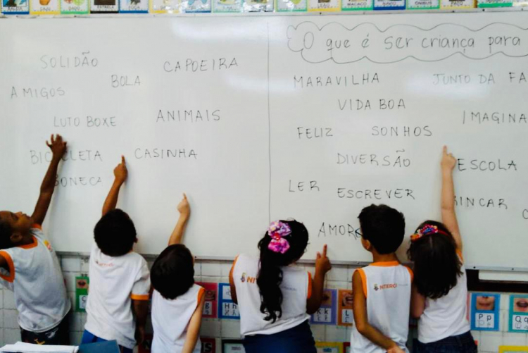 Seis crianças estão de costas para a câmera e de frente para um quadro branco com palavras escritas