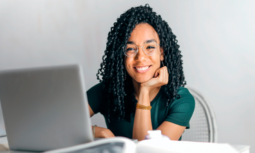 Mulher negra de óculos sorrindo sentada em frente a mesa com laptop
