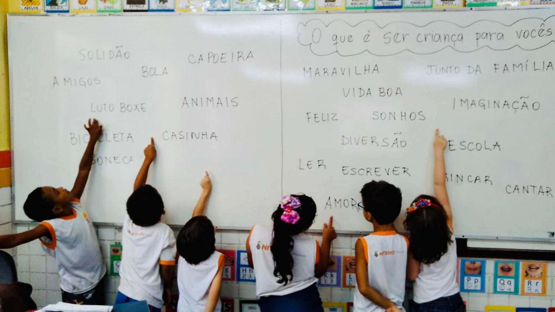 Seis crianças estão de costas para a câmera e de frente para um quadro branco com palavras escritas