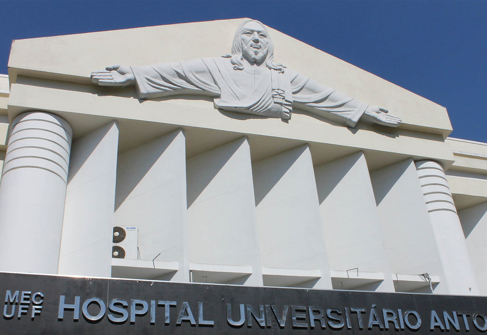 Foto da fachada do Hospital Universitário Antônio Pedro, onde se lê o nome da unidade e a escultura localizada no topo da edificação.