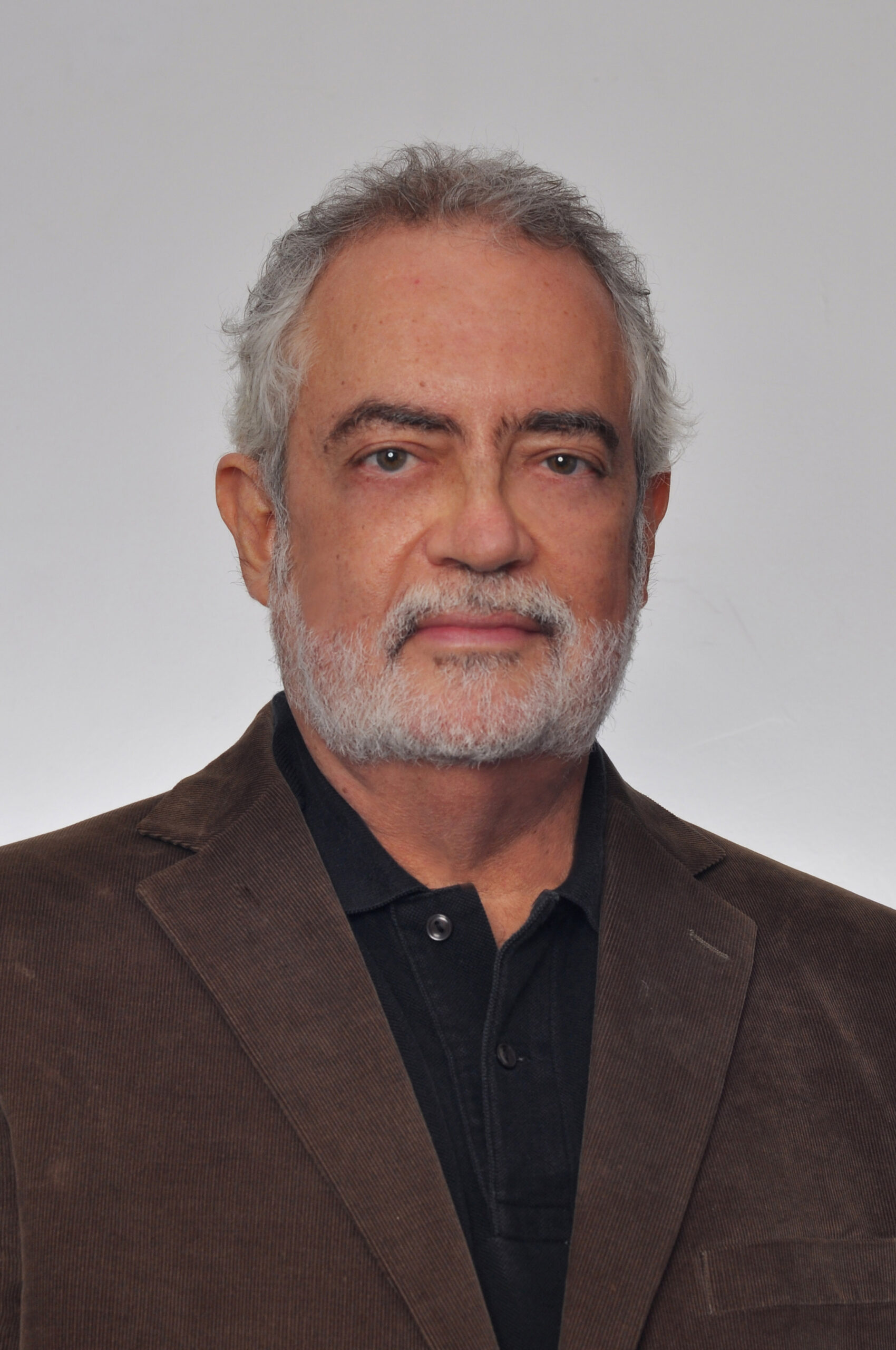 Foto de homem branco de barba e cabelos grisalhos, vestindo terno marrom e blusa preta.