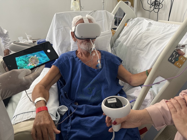 foto de paciente idoso, sentado no leito do hospital, utilizando um óculos de realidade virtual branco. Ao lado aparece uma mão segurando um tablet com imagem do fundo do mar.